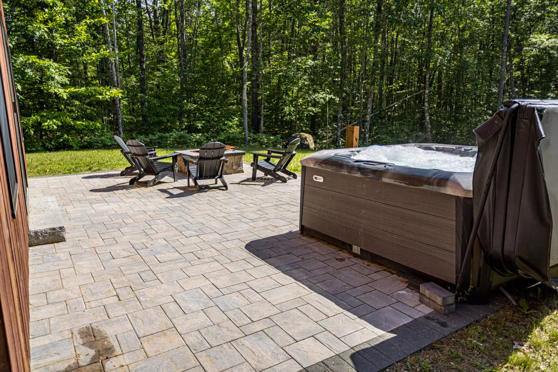 Paver patio with three Adirondack chairs around a fire pit, with a covered hot tub at right, edged by tall woods
